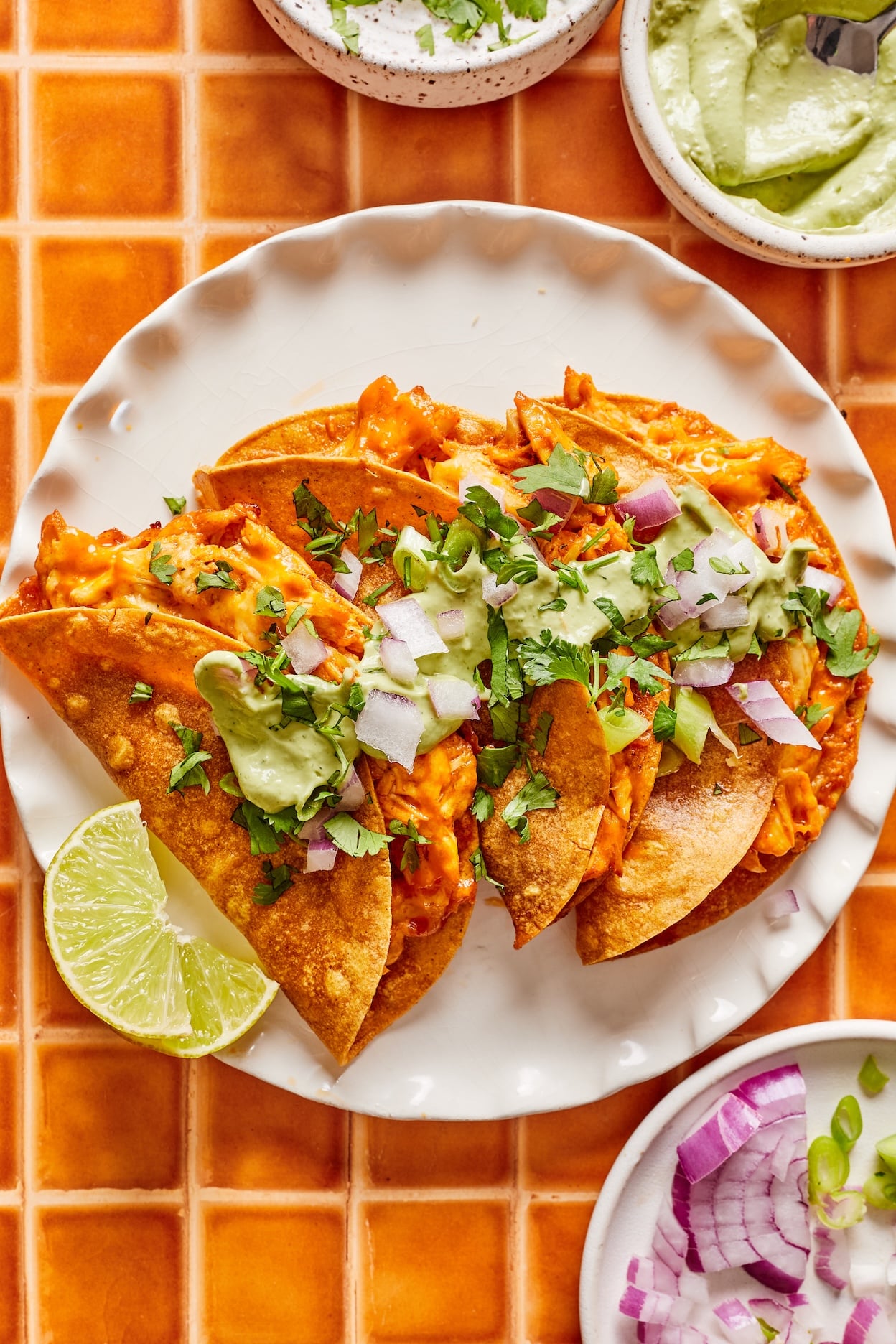 Crispy buffalo chicken tacos arranged on a white plate, garnished with cilantro, chopped red onion, and avocado cilantro sauce, with lime wedges and a bowl of additional chopped red onion, cilantro, and avocado cilantro sauce alongside.