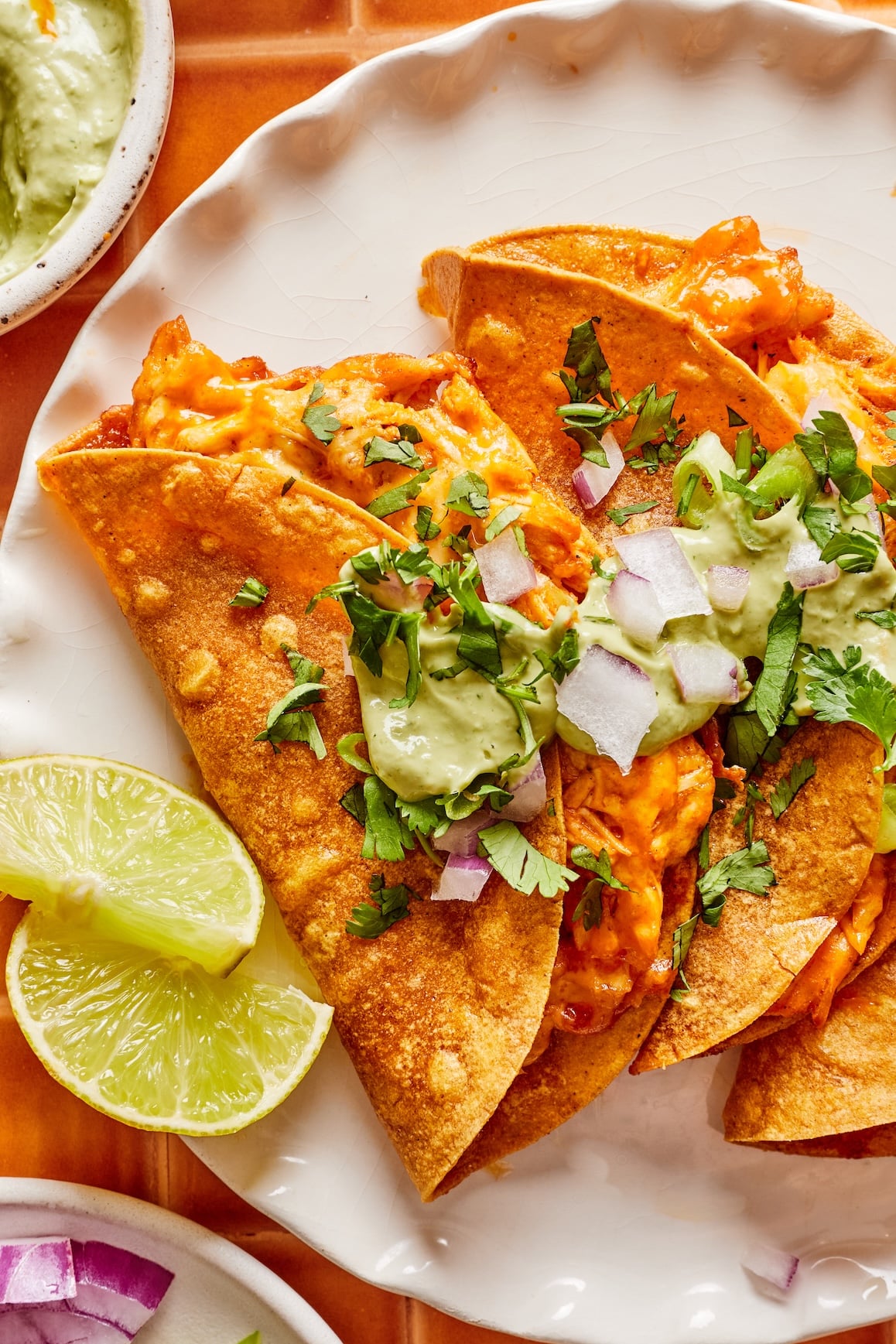 Close-up of crispy buffalo chicken tacos on a plate, garnished with cilantro, chopped red onion, and avocado cilantro sauce, with lime wedges on the side.