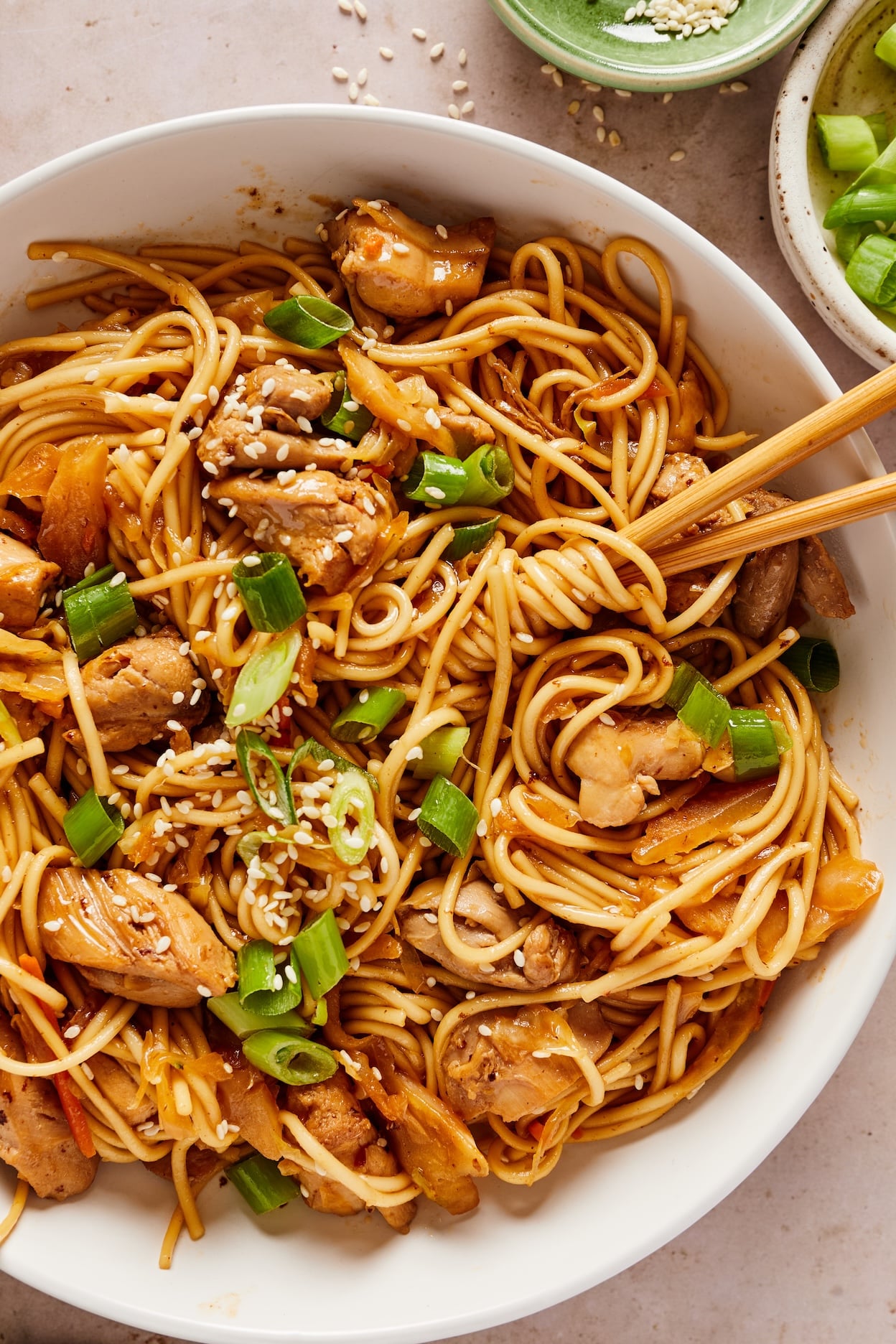 Bowl of honey garlic chicken noodles garnished with sliced green onions and sesame seeds, with chopsticks holding a bundle of noodles.