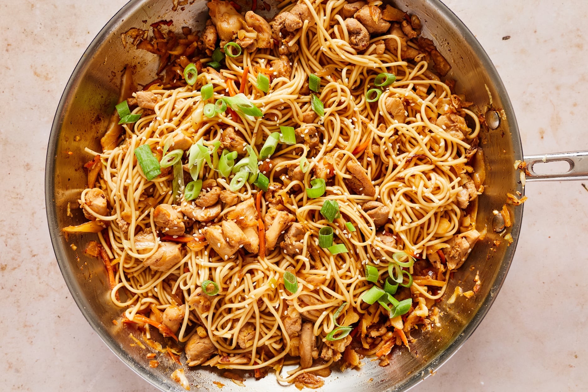 Honey garlic chicken, noodles, and cabbage tossed with sauce in a large skillet until evenly coated, garnished with green onions and sesame seeds.
