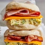 Two freezer breakfast sandwiches stacked on parchment paper, each with a square of baked eggs studded with broccoli, a slice of cheddar, and ham on the top sandwich and bacon on the bottom, on English muffins