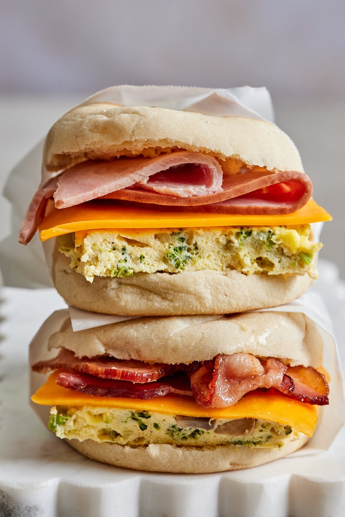 Two freezer breakfast sandwiches stacked on parchment paper, each with a square of baked eggs studded with broccoli, a slice of cheddar, and ham on the top sandwich and bacon on the bottom, on English muffins