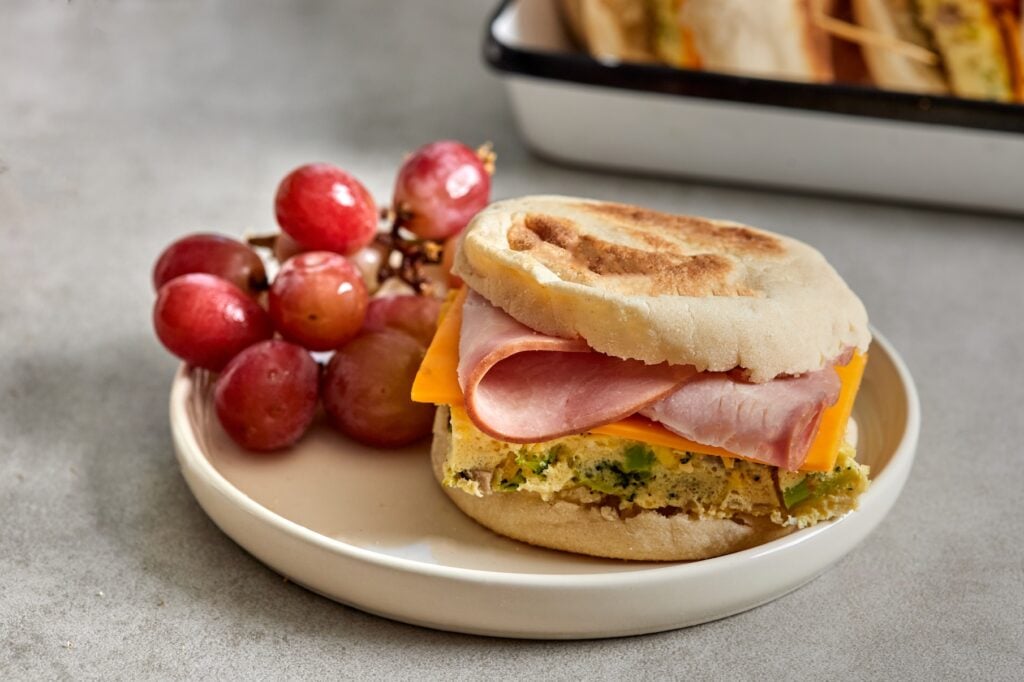 One ham breakfast sandwich on a white plate next to a handful of red grapes.