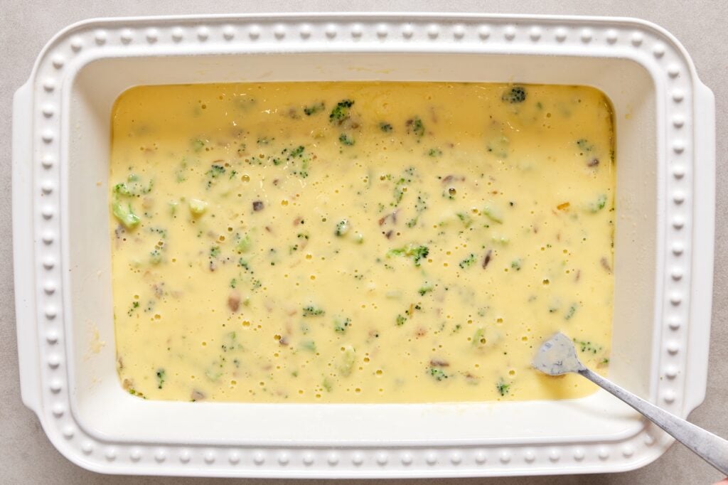 A white baking dish with veggies at the bottom and the egg mixture poured over the top before going in the oven.