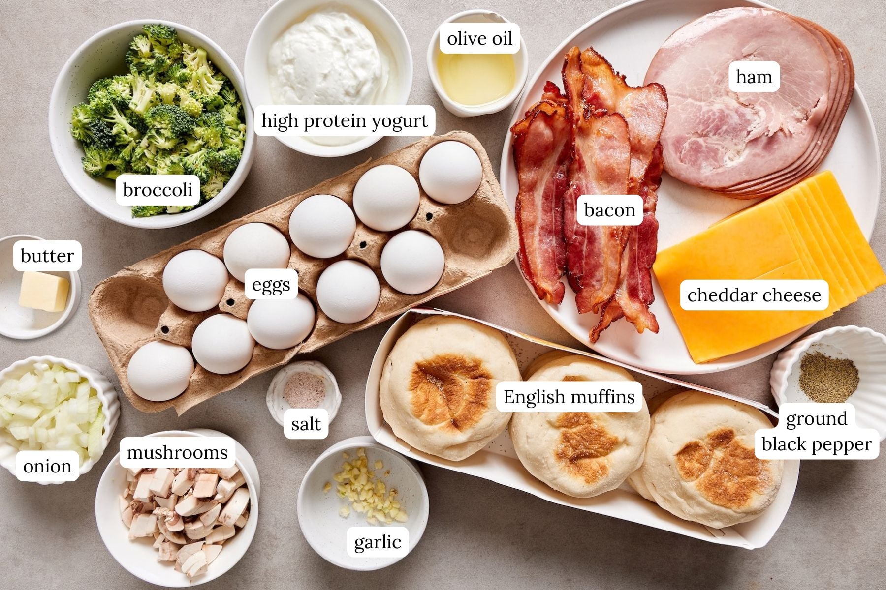 Ingredients for veggie freezer breakfast sandwiches on a counter: eggs, high protein yogurt, chopped broccoli, mushrooms, diced onion, garlic, English muffins, cheddar slices, ham, bacon, butter, olive oil, salt, and pepper
