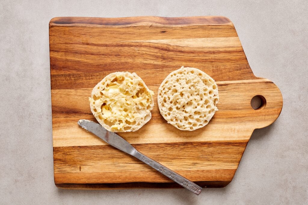 An open face english muffin on a cutting board next to a butter knife. One side of the english muffin has butter spread across.
