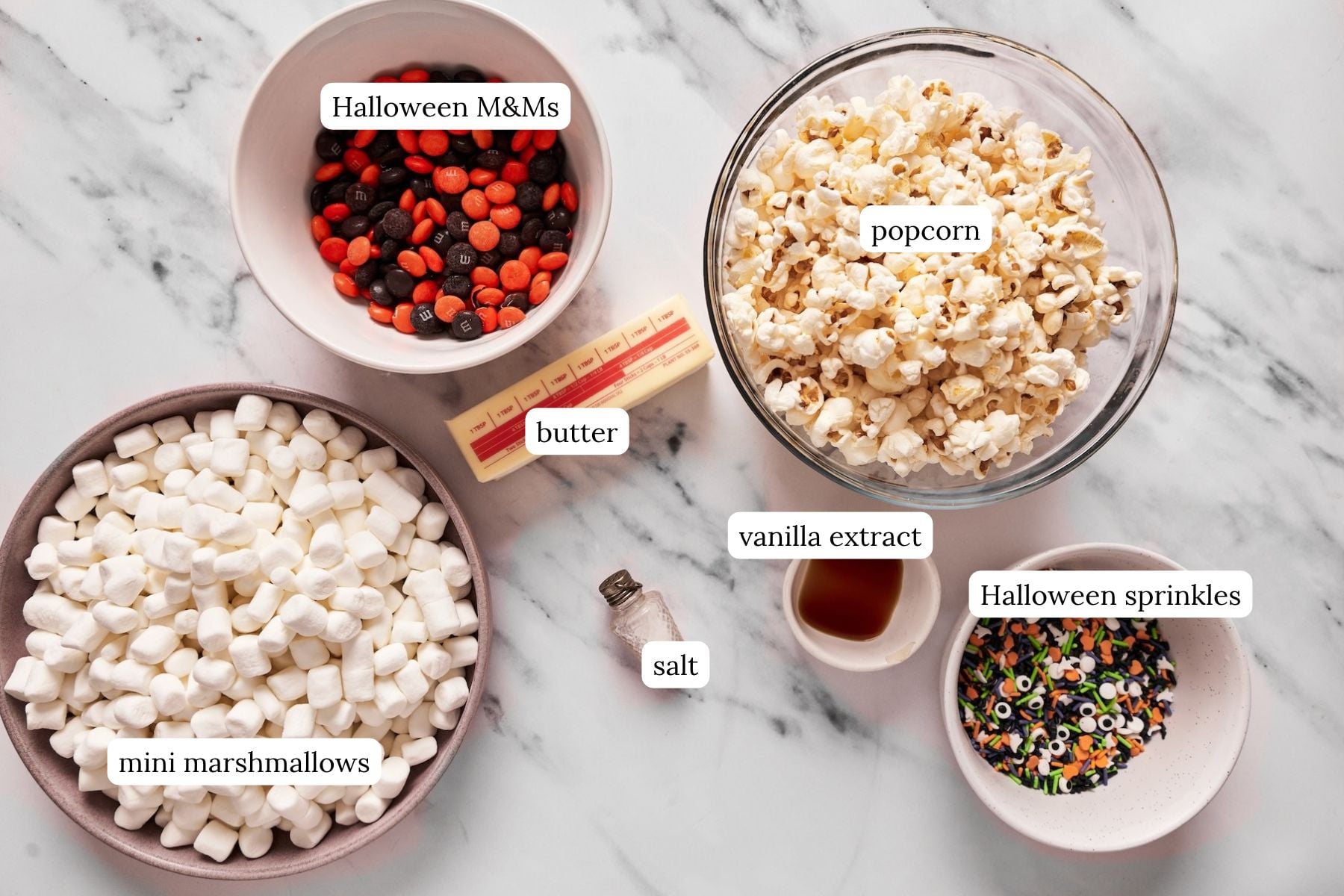 Labeled ingredients for halloween marshmallow popcorn balls on a marble countertop.