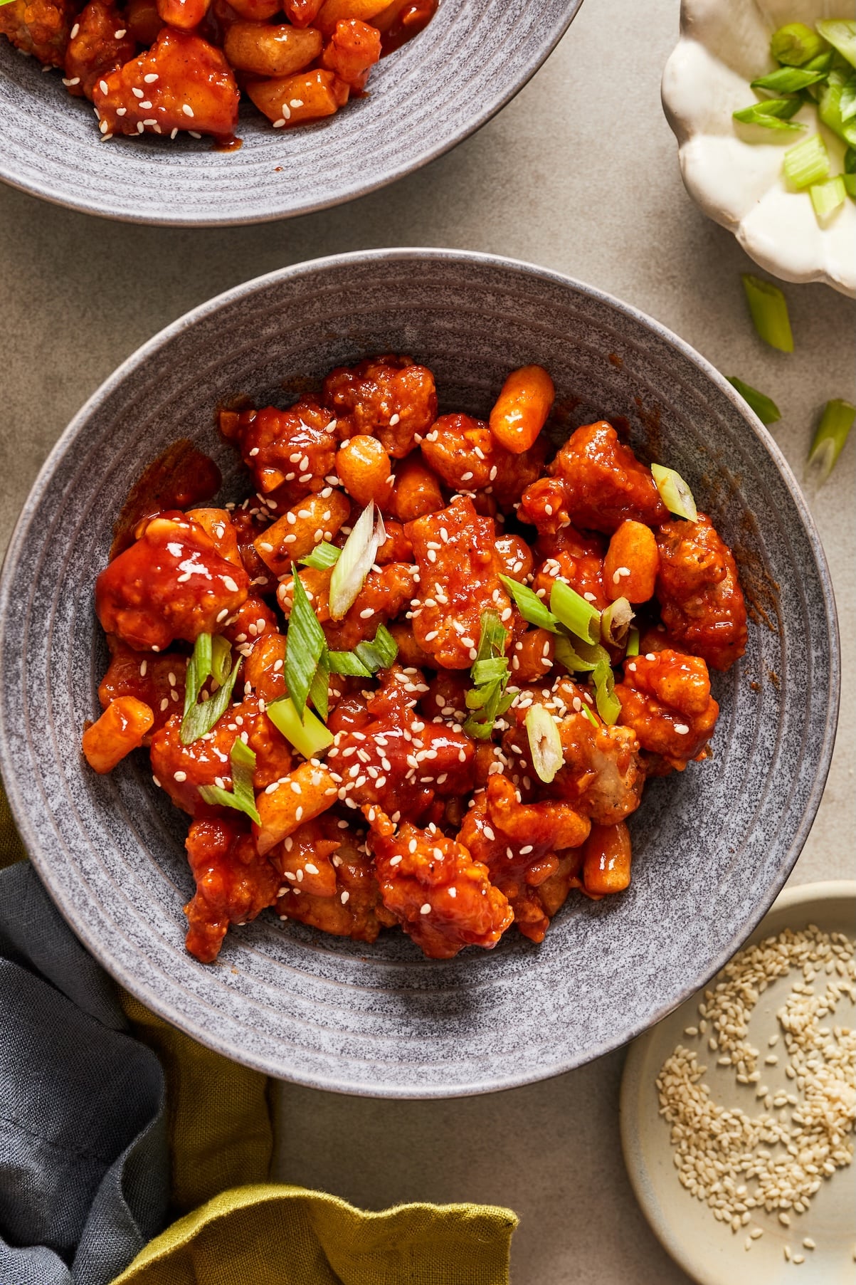 An overhead shot of Dakgangjeong tossed in a sweet and spicy sauce in a gray bowl topped with sesame seeds and green onions.