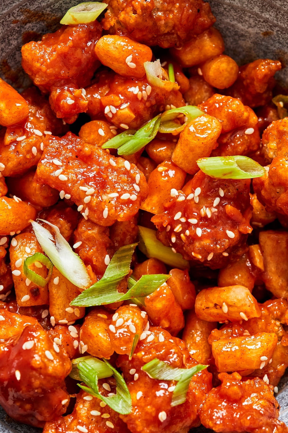 A close up shot of Dakgangjeong tossed in a sweet and spicy sauce in topped with sesame seeds and green onions.