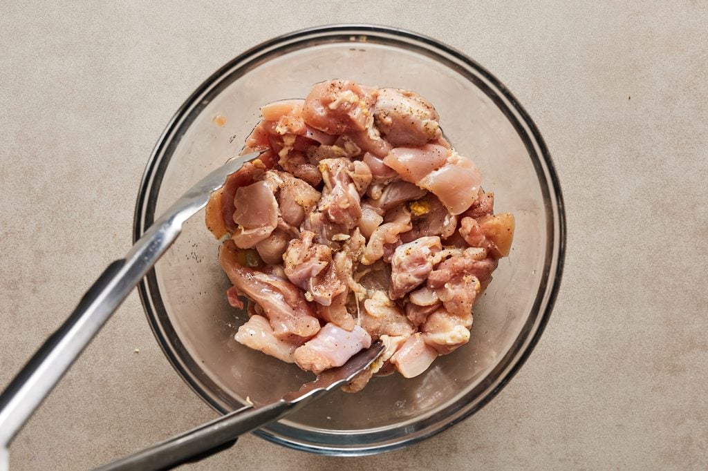 Raw chicken thighs marinating in a glass bowls with tongs.